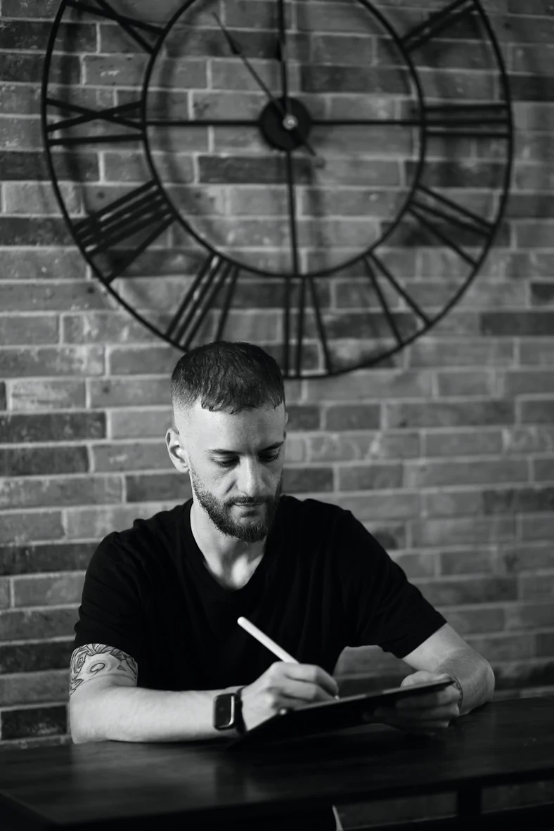 Ritratto in bianco e nero di Alessio Visentin che disegna su un tablet, seduto a un tavolo davanti a un muro di mattoni con un grande orologio a numeri romani.