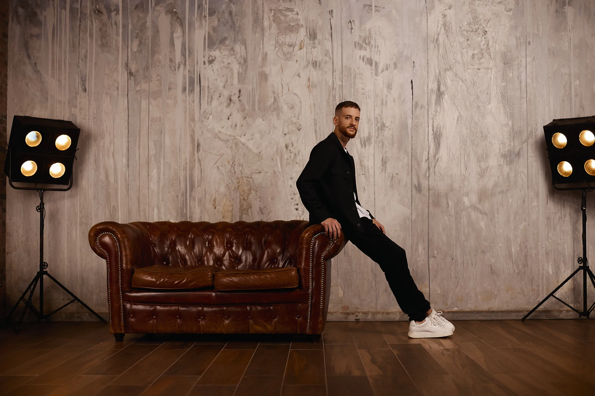 Alessio Visentin appoggiato allo schienale di un divano Chesterfield in pelle marrone, in uno studio dal muro di cemento grezzo, con due fari da set accesi ai lati.
